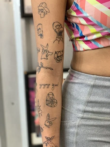 A person's arm with multiple tattoos, including a skull, a skull with a hat, a rabbit, a skull with sunglasses, a lightning bolt, the phrase '777 good!', a spider web, a bunny, and a flying rabbit.