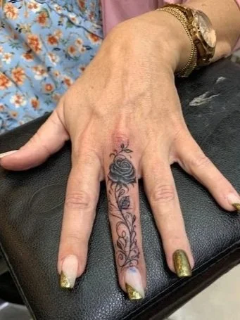 A hand with gold-painted nails resting on a black cushion, showing a tattoo of a rosette and floral design between the fingers.