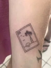 Tattoo of a simple black and white postage stamp with a palm tree and sunset scene on the person's forearm.