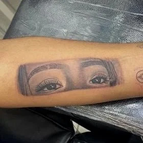 Tattooed arm with the year '1975' at the top, a monochrome eye portrait, an outline of a person holding a child, and small text.