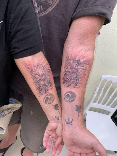 Two people showing their tattooed forearms. The tattoos include a dragon and a tiger, with additional smaller tattoos of a moon, palm trees, and a crown.