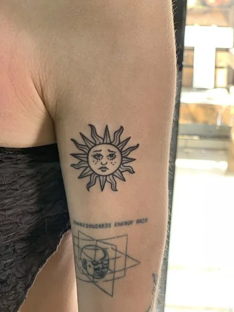 Tattoo of a sun with a human face on upper arm, with a geometric design and face on surrounding elements below.