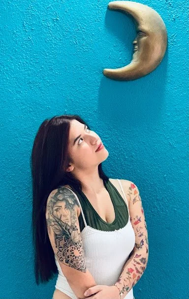 A woman with long dark hair and tattoos on her arms looking up at a crescent moon decoration on a blue wall.