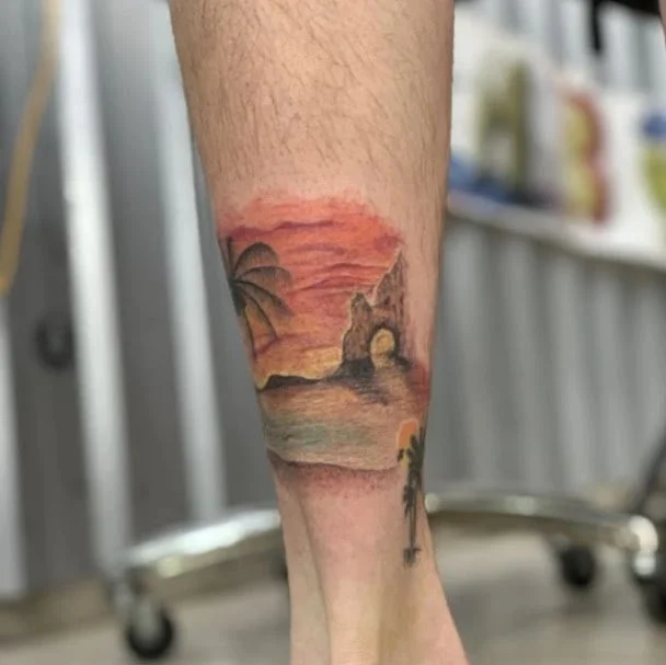 Tattoo of a beach sunset with palm trees, rocky arch, and waves on a person's lower leg.