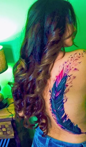 Side view of a woman with long, wavy brown hair and a colorful feather tattoo on her back.