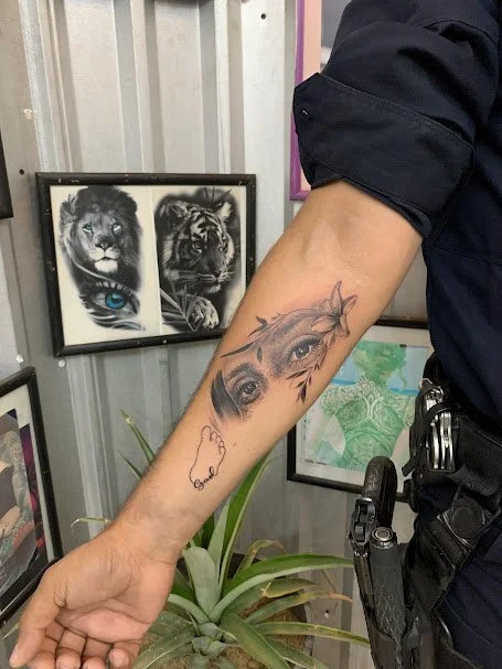 A person's arm with a tattoo of realistic eyes and a branch with leaves. In the background, there are framed pictures of a lion, a tiger, and a peacock feather.