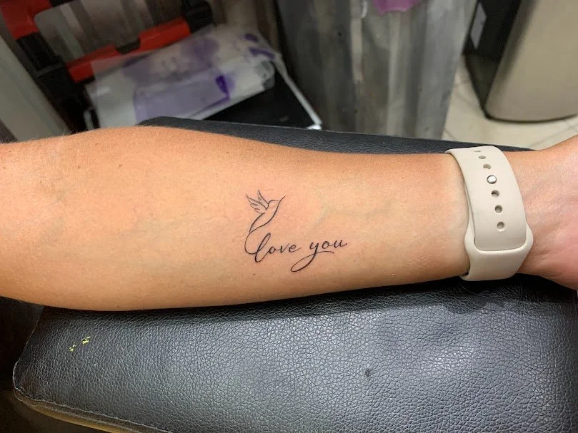 Tattoo of a hummingbird and the words 'love you' on a person's forearm.