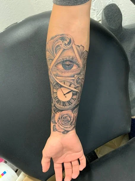 Tattoo on forearm depicting an eye within swirling clouds, a clock with Roman numerals, and a rose at the bottom.