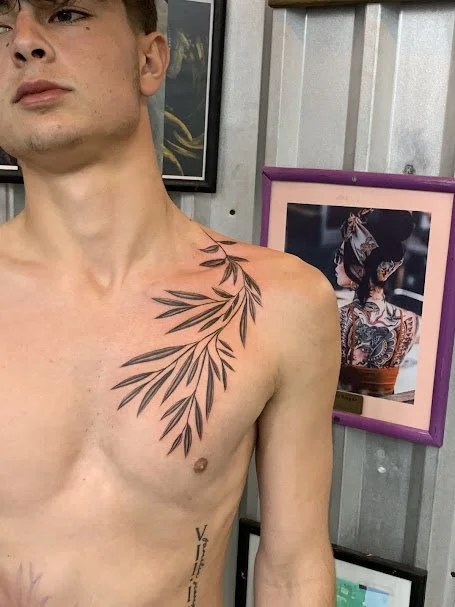 A young man with a tattoo of leaves on his right shoulder and chest standing in front of framed artwork in an indoor setting.
