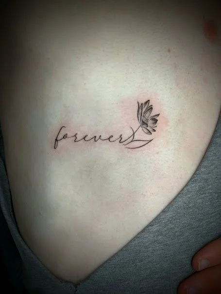 Tattoo of the word 'forever' in cursive with a small flower and stem on the skin.