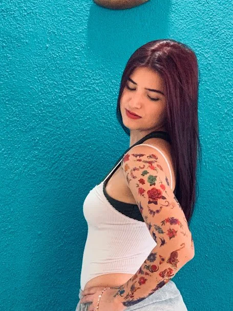 A woman with long dark red hair standing against a bright teal wall, wearing a white tank top with black straps and showing a tattooed arm.