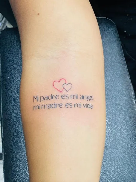 Tattoo of two intertwined hearts, one red and one black, above the Spanish text "Mi padre es mi ángel. mi madre es mi vida" on a person's arm.