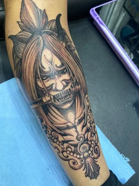 Tattoo of a fierce wolf woman with long hair and a bone in her mouth, detailed shading, and ornamental elements on the arm.