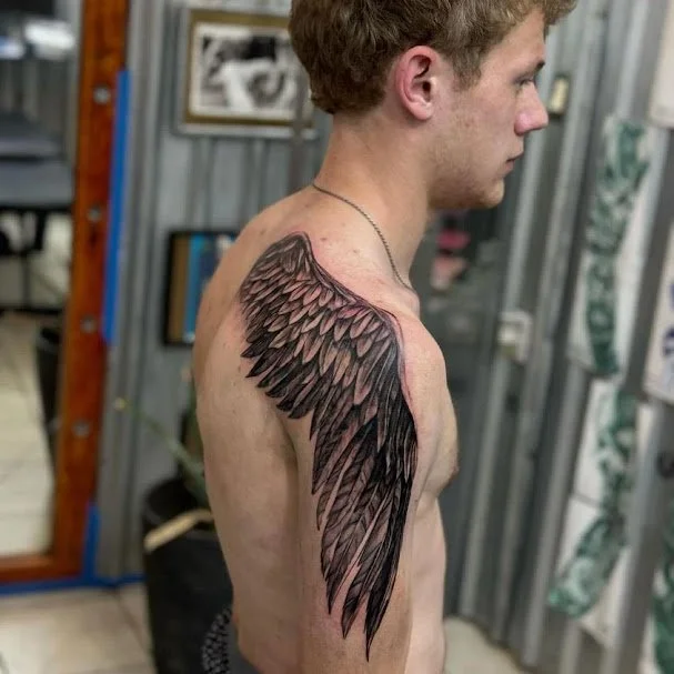 A young man with a large black and gray wing tattoo on his shoulder and upper arm, standing sideways in a tattoo studio.