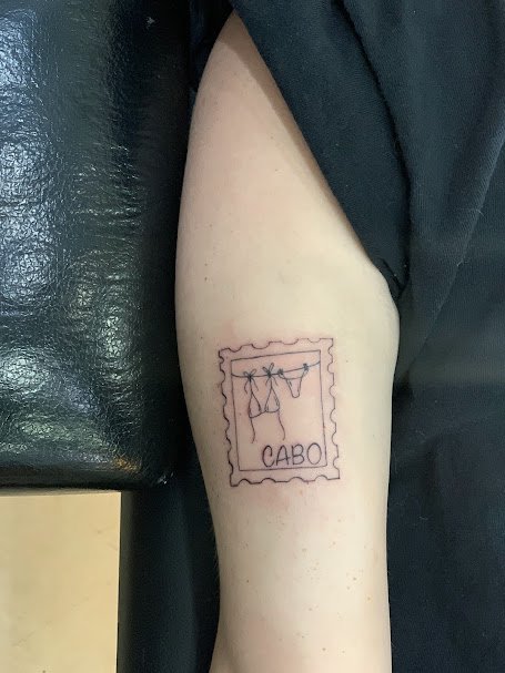 Tattoo of a postage stamp with a hanging clothesline and the word 'Cabo' on a person's upper arm.