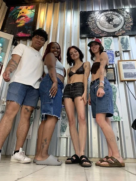 Four young women and one young man standing together indoors, smiling for the camera, with colorful artwork and framed pictures on the wall behind them.
