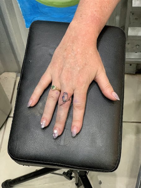 A person's left hand with a small blue bird tattoo on the ring finger, resting on a black padded surface. The person is wearing a gold ring with a green stone on the ring finger, and has painted nails with white and pink designs.