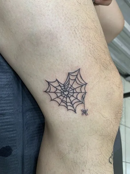 Tattoo of a spider web with a small spider hanging from it on a person's upper thigh.