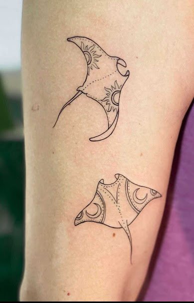 Two tattoos of manta rays with sun, moon, and star motifs on a person's arm.
