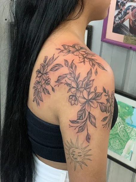 Tattoo of floral branches and a smiling sun on a woman's upper arm and shoulder.