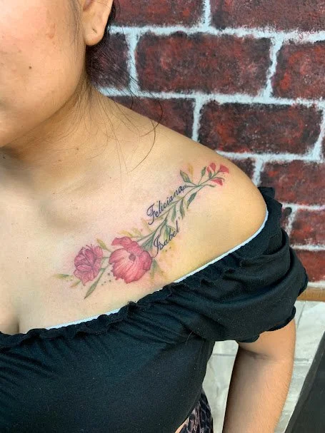 Tattoo of pink flowers with green leaves and the names 'Felicia' and 'Talia' on the person's shoulder near a red brick wall.