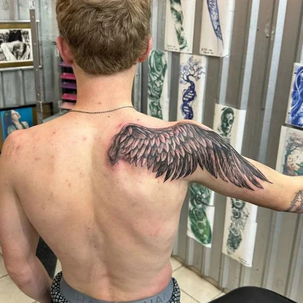 A person with short blond hair, visible back and shoulders, displaying a large wing tattoo on their right shoulder and upper arm. The tattoo has detailed feathers. The background shows tattoo designs and artwork on the wall.
