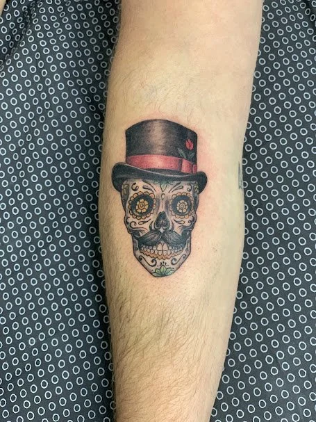 Tattoo of a decorated sugar skull wearing a black top hat with a red band and a small red heart.