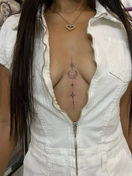 A woman wearing a white zip-up jumpsuit with a plunging neckline, revealing a chest tattoo of star and moon designs, and wearing a gold necklace with a heart-shaped pendant.