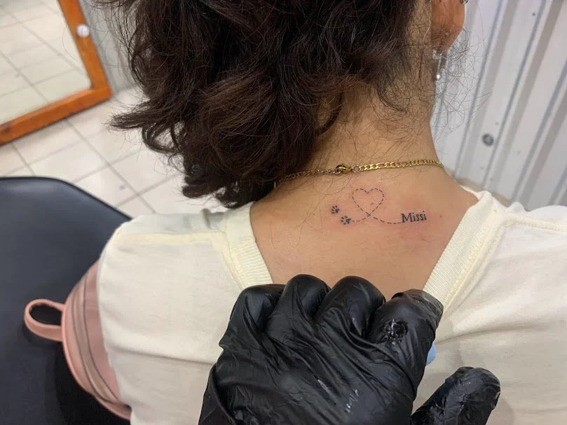 Tattoo on the back of a woman's neck showing a heart shape made of dots, a dotted line leading to the name 'Missi', and a paw print.