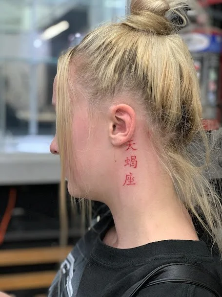 A woman with blonde hair tied up in a messy bun, showing red Chinese characters tattooed on her neck in profile view.