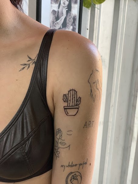 Tattooed shoulder and upper arm of a woman wearing a black sleeveless top, with tattoos including a cactus in a pot, a face with closed eyes and a flower, and the words 'ART' and 'say whatever you want'.
