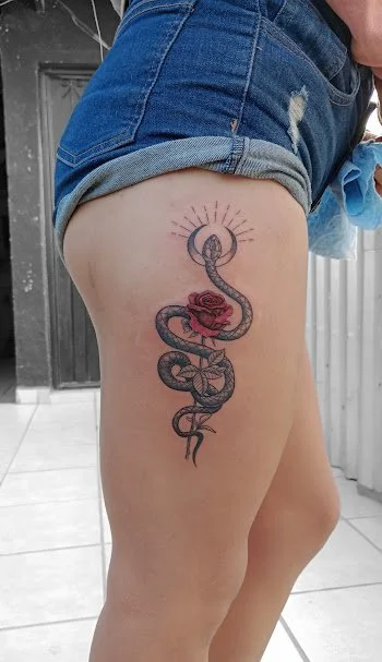 Tattoo of a snake entwined around a red rose on a person's thigh, with rays emanating from the top of the design.