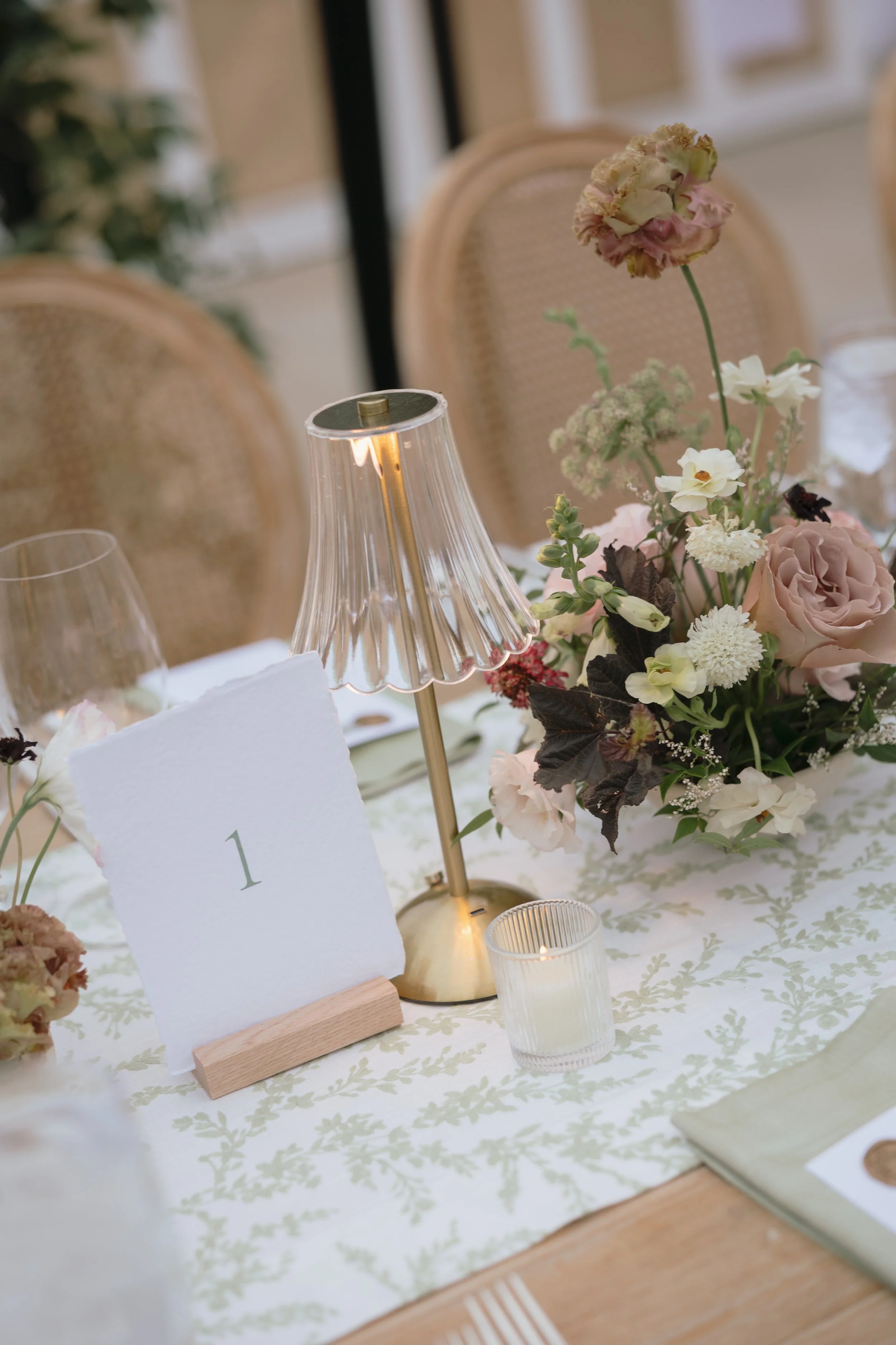 Elegant table setting with a floral centerpiece, a table number card, a glass candle holder, a small candle, and a table lamp, all on a decorated tablecloth.