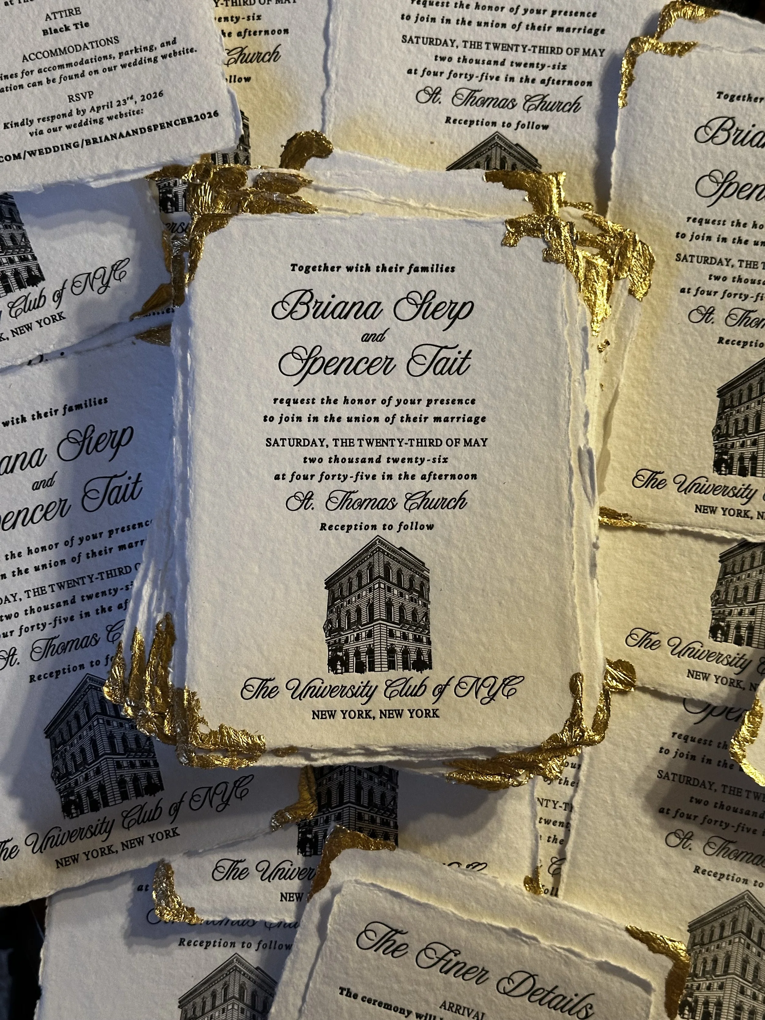 Wedding invitation cards with black text and a black illustration of a building, decorated with gold foil accents on the torn edges, arranged layered on a surface.
