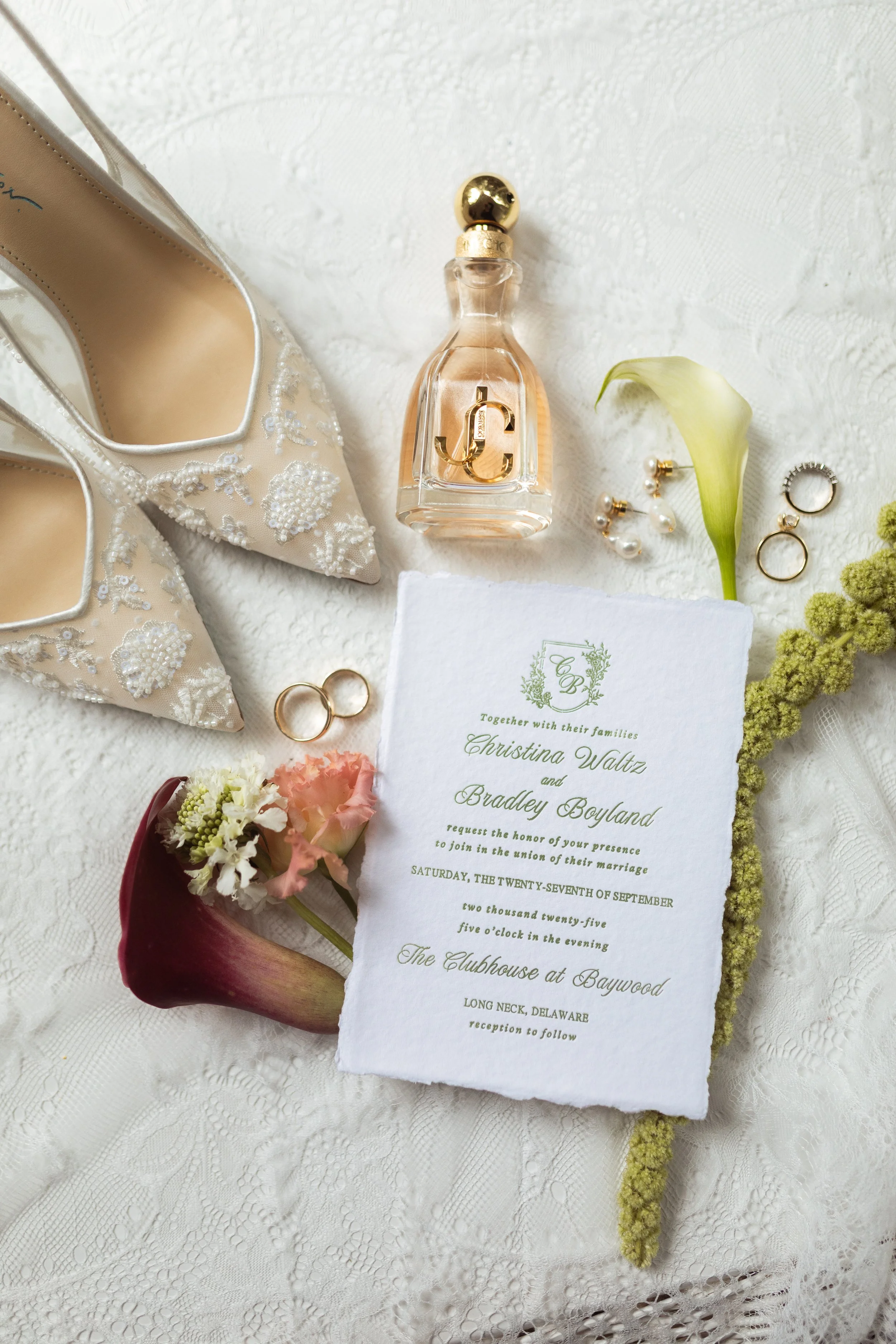 Wedding invitation with white lace tablecloth, cream embroidered high heels, perfume bottle, gold wedding rings, pearl earrings, a calla lily, a pink and white flower, and a green flowering branch.