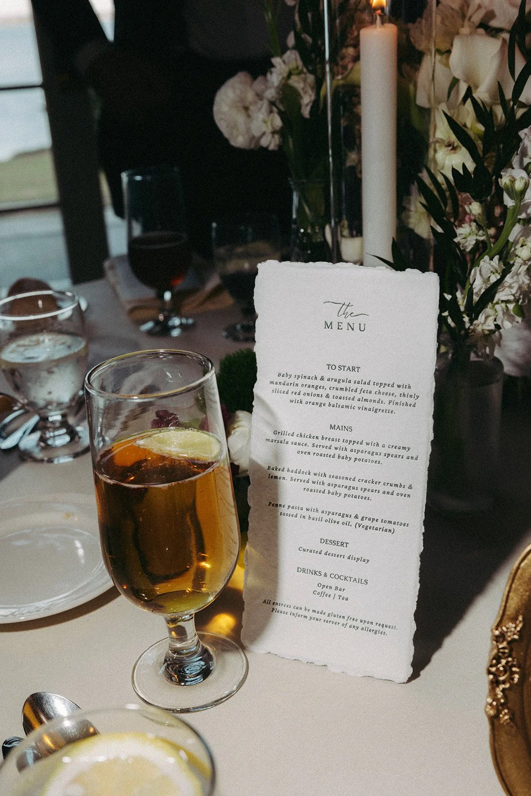 A table setting at a formal event with a cocktail glass filled with a drink and a lemon wedge, a paper menu card, water glasses, and floral arrangements in the background.