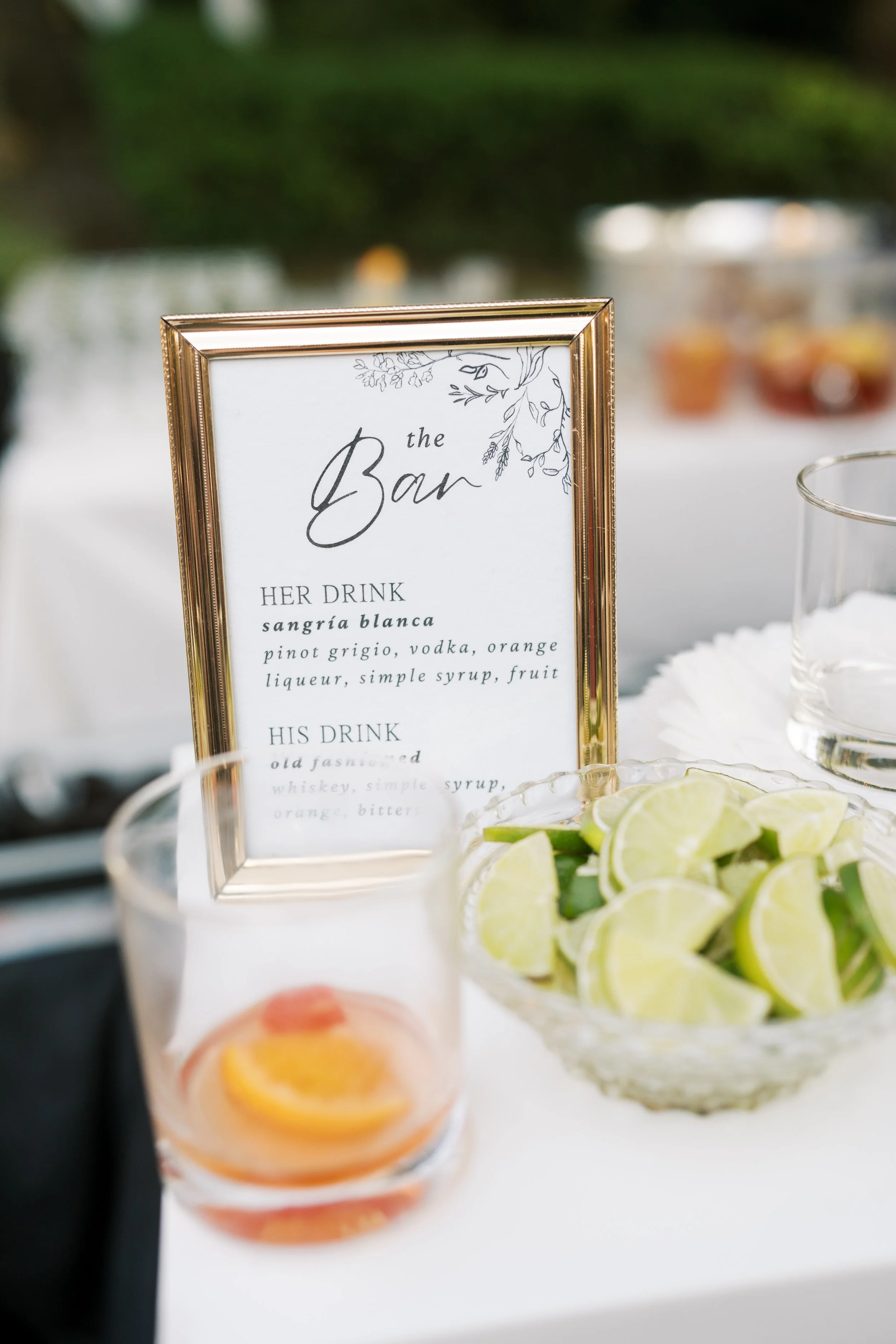 A framed sign at a bar station listing drinks for women and men, with a bowl of lime wedges and a glass of a pink cocktail with citrus slices.