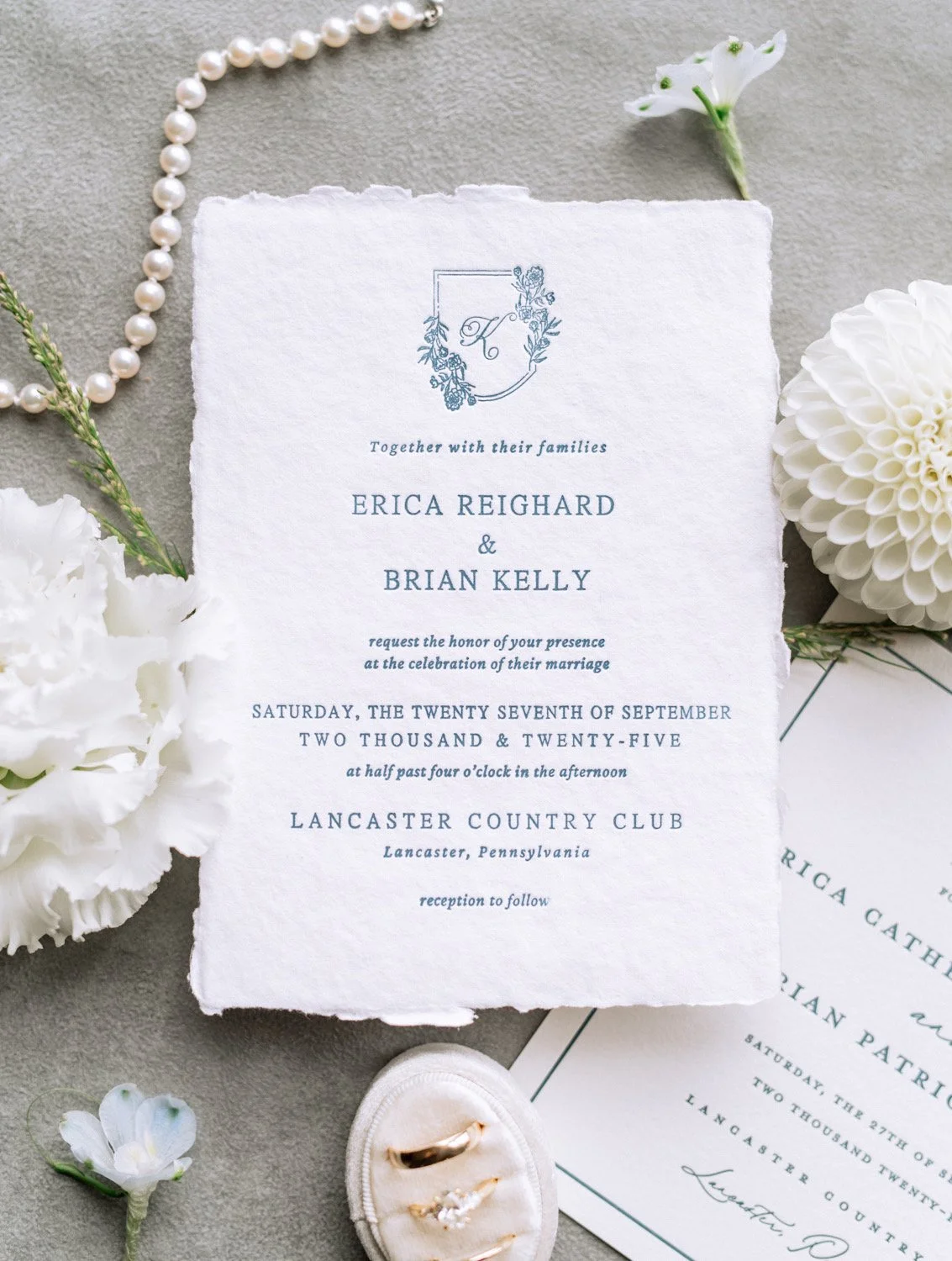 Wedding invitation surrounded by white flowers, pearl necklace, and wedding rings on a jewelry box.