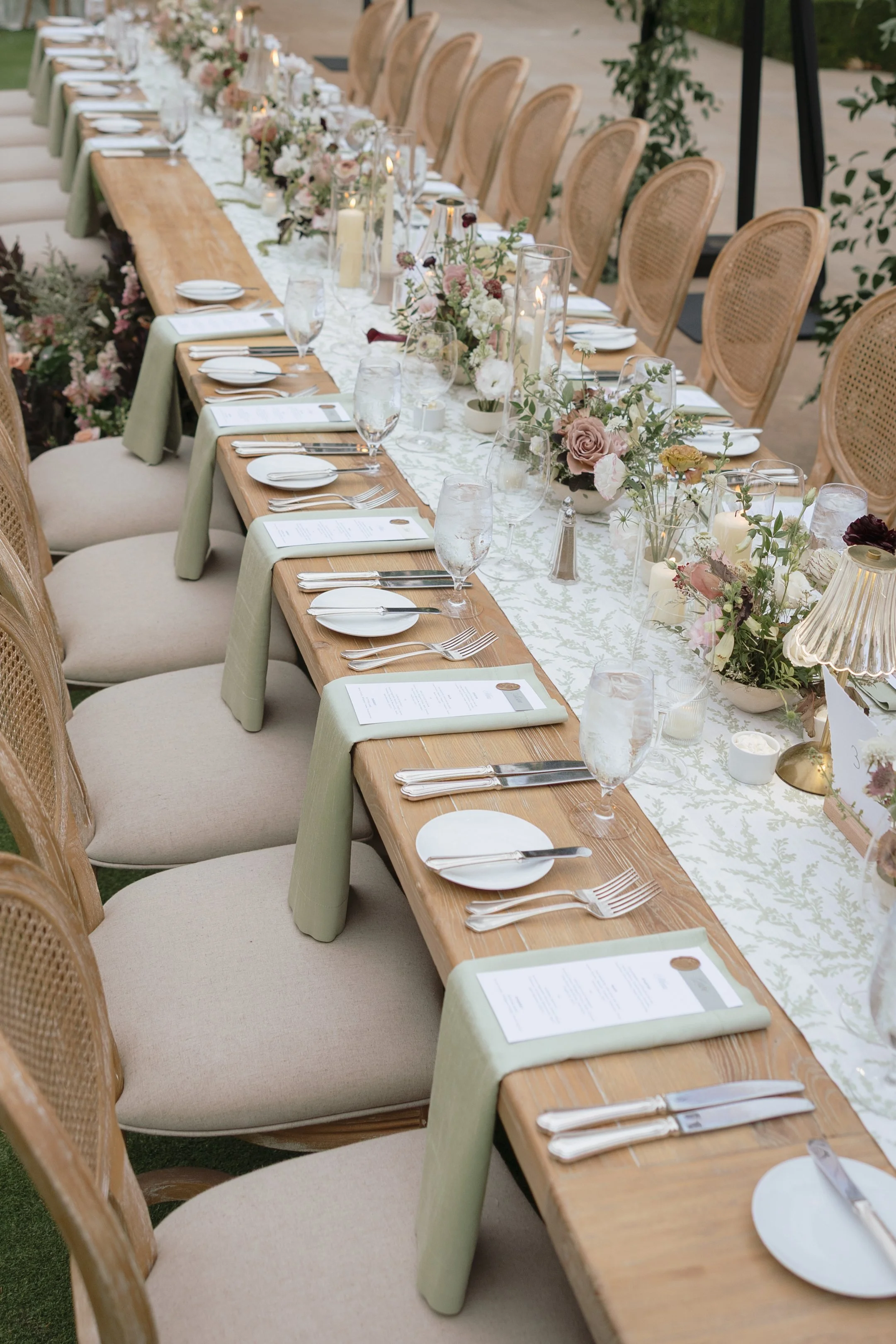 Long wooden banquet table set for a formal event with floral centerpieces, candles, and neatly arranged glassware and silverware, surrounded by woven rattan chairs.