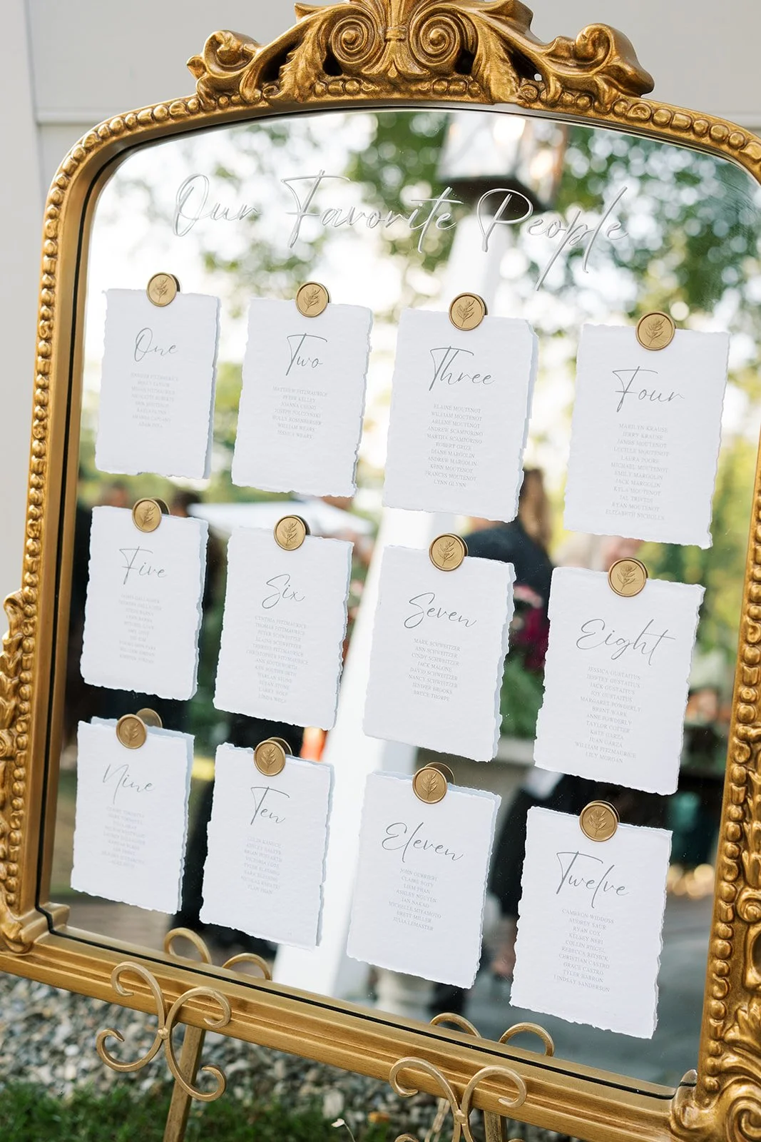 A decorative gold-framed mirror with handwritten seating chart cards pinned to the glass. The cards are numbered from one to twelve with guests' names listed below each number, used as a table seating chart for an event.