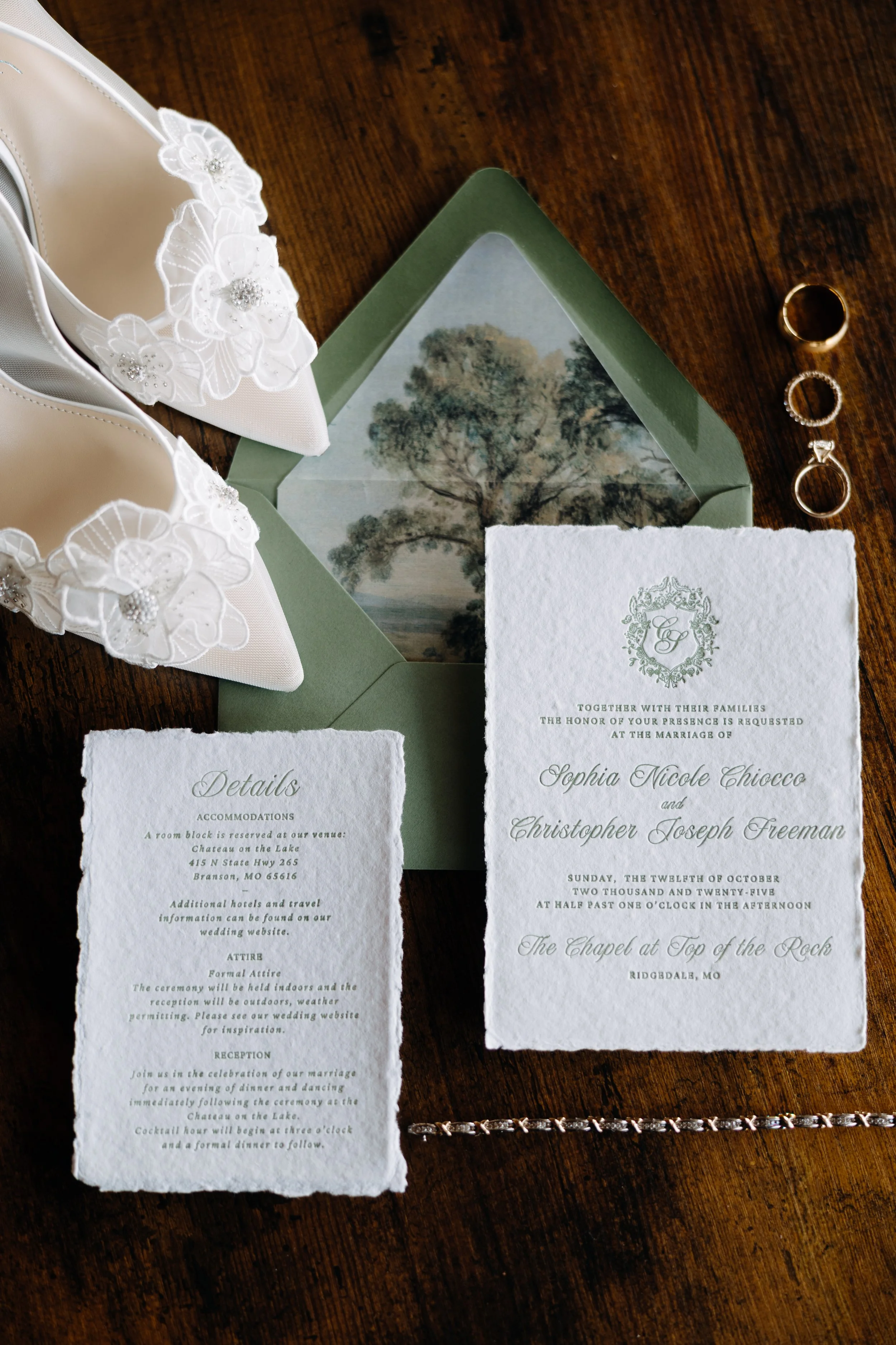 Wedding invitation, white high heels with floral embellishments, wedding rings, and a decorative envelope with a tree illustration inside, all arranged on a wooden surface.
