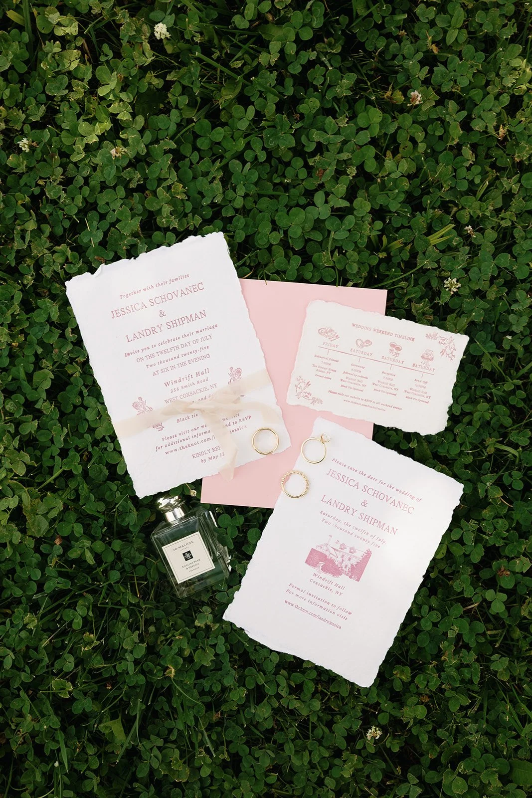 Wedding invitation suite, including two main invitation cards, a smaller event schedule card, and some rings, all placed on a bed of green clover with a pink envelope and a perfume bottle nearby.