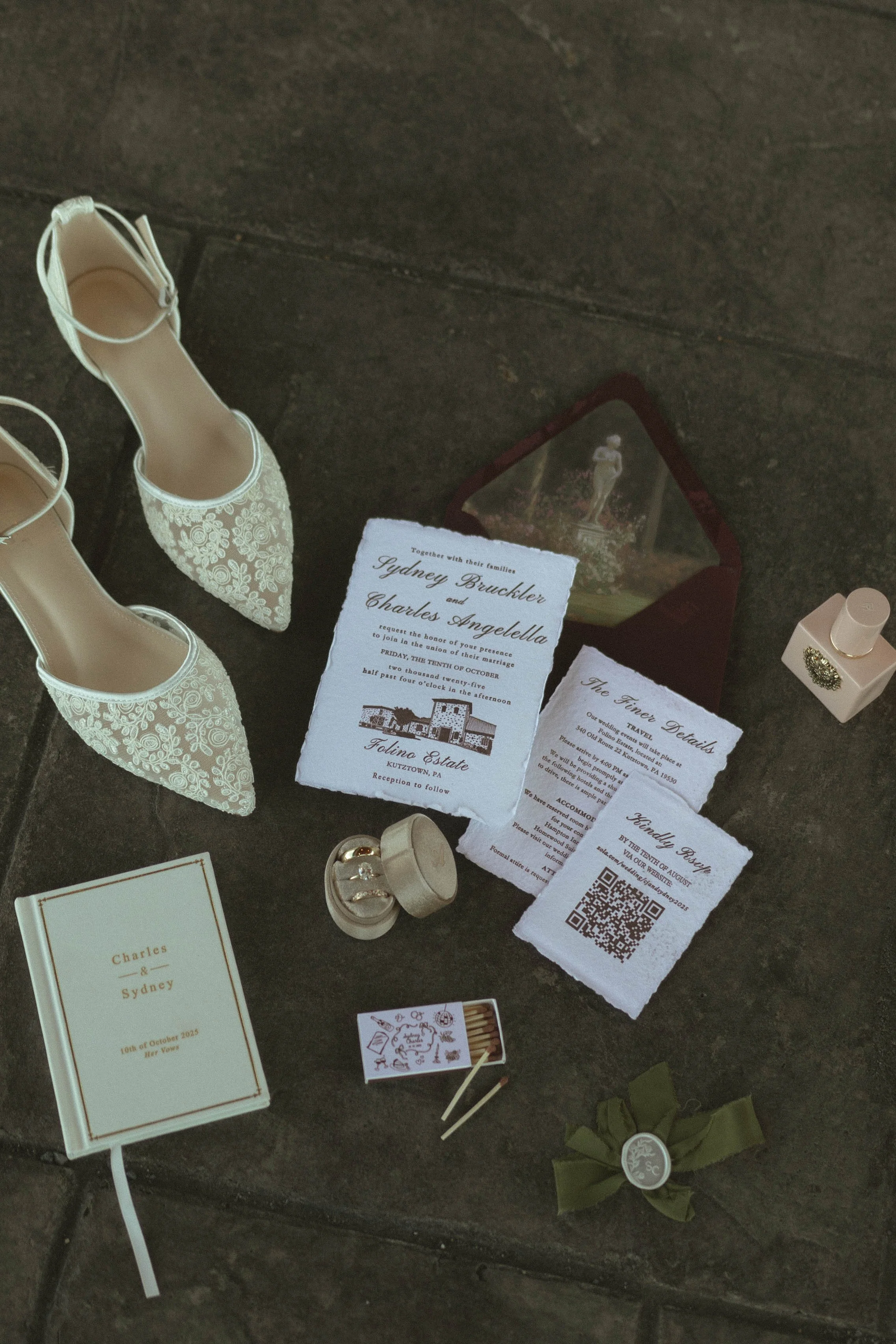 Wedding invitation suite with white lace shoes, rings, wedding details, and a monogrammed green ribbon, all displayed on a dark stone surface.