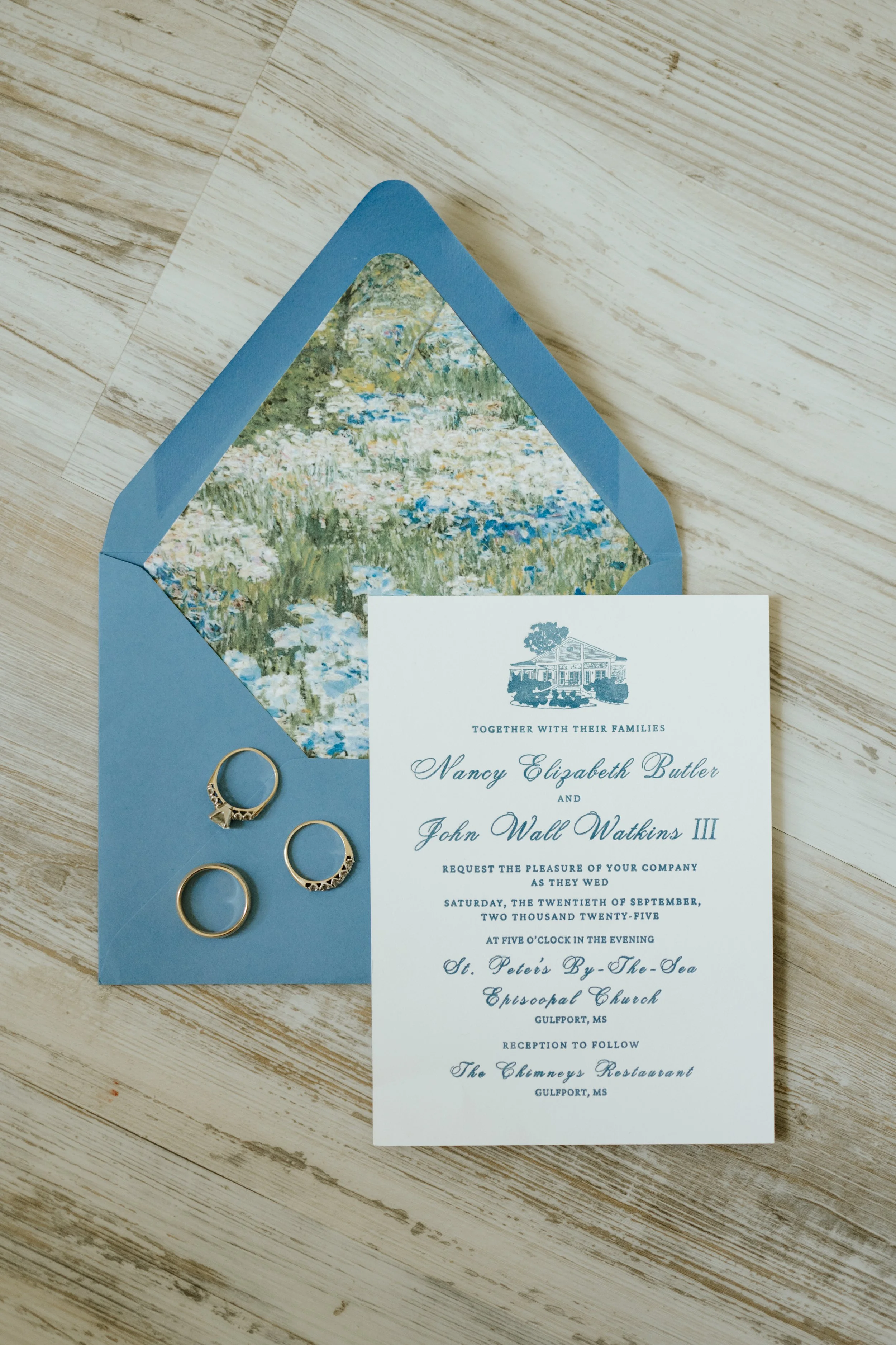 Wedding invitation card placed on a wooden surface, partially inside a blue envelope lined with Impressionist painting design, with three gold rings beside it.