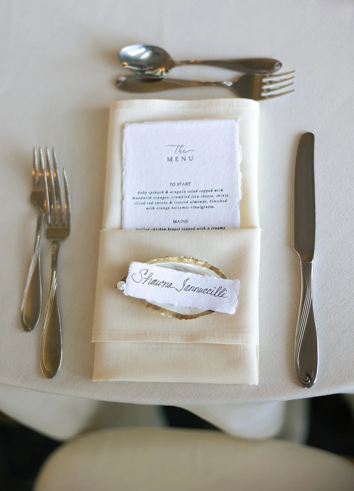 Place setting with a white plate with an oyster shell and a name tag reading 'Shawna Sannucilli,' a folded cream napkin, a printed menu, a knife on the right, a dinner fork on the left, and a dessert spoon and fork at the top.