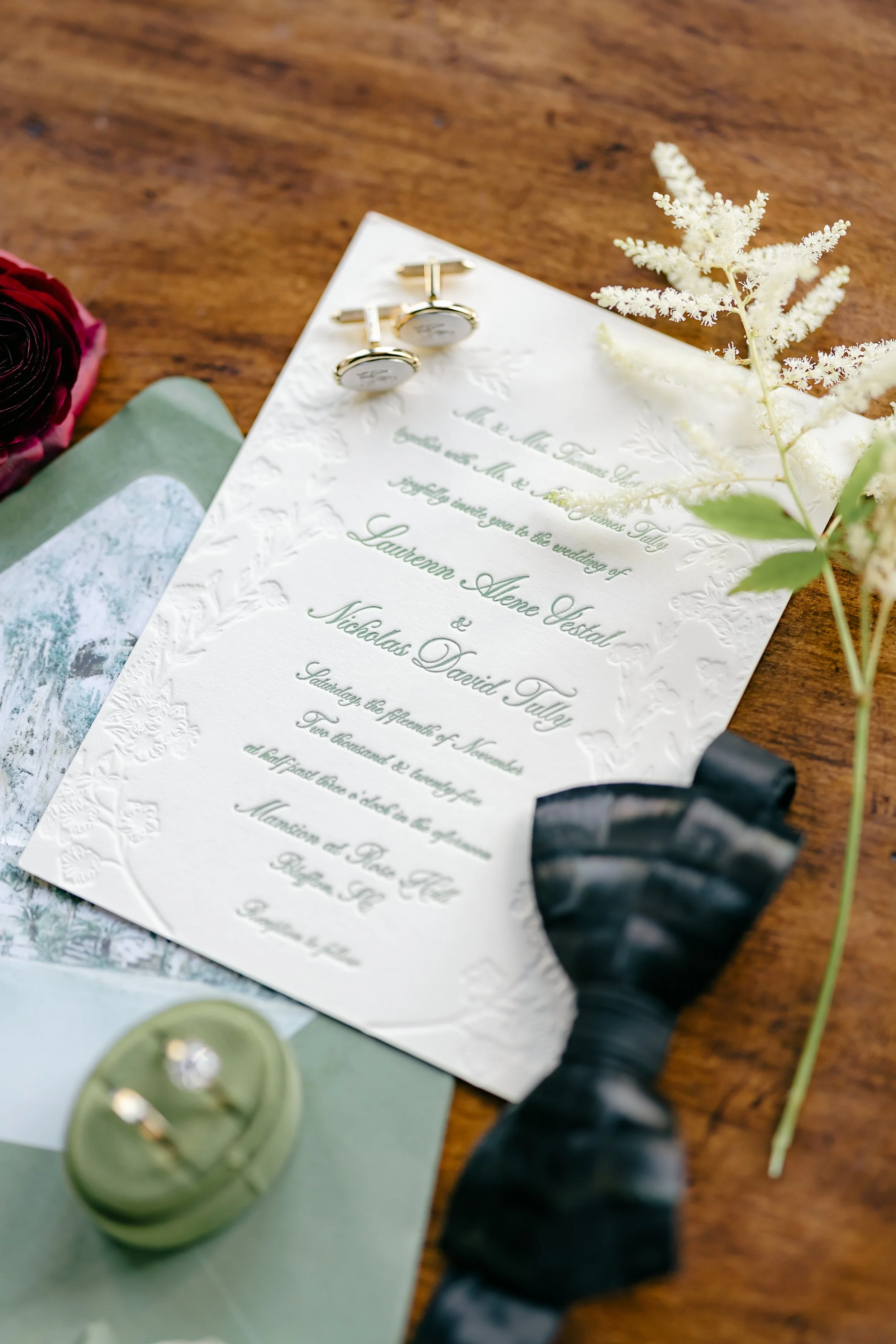 Wedding invitation on a wooden surface with a pair of silver cufflinks, a white flower, and a black ribbon.