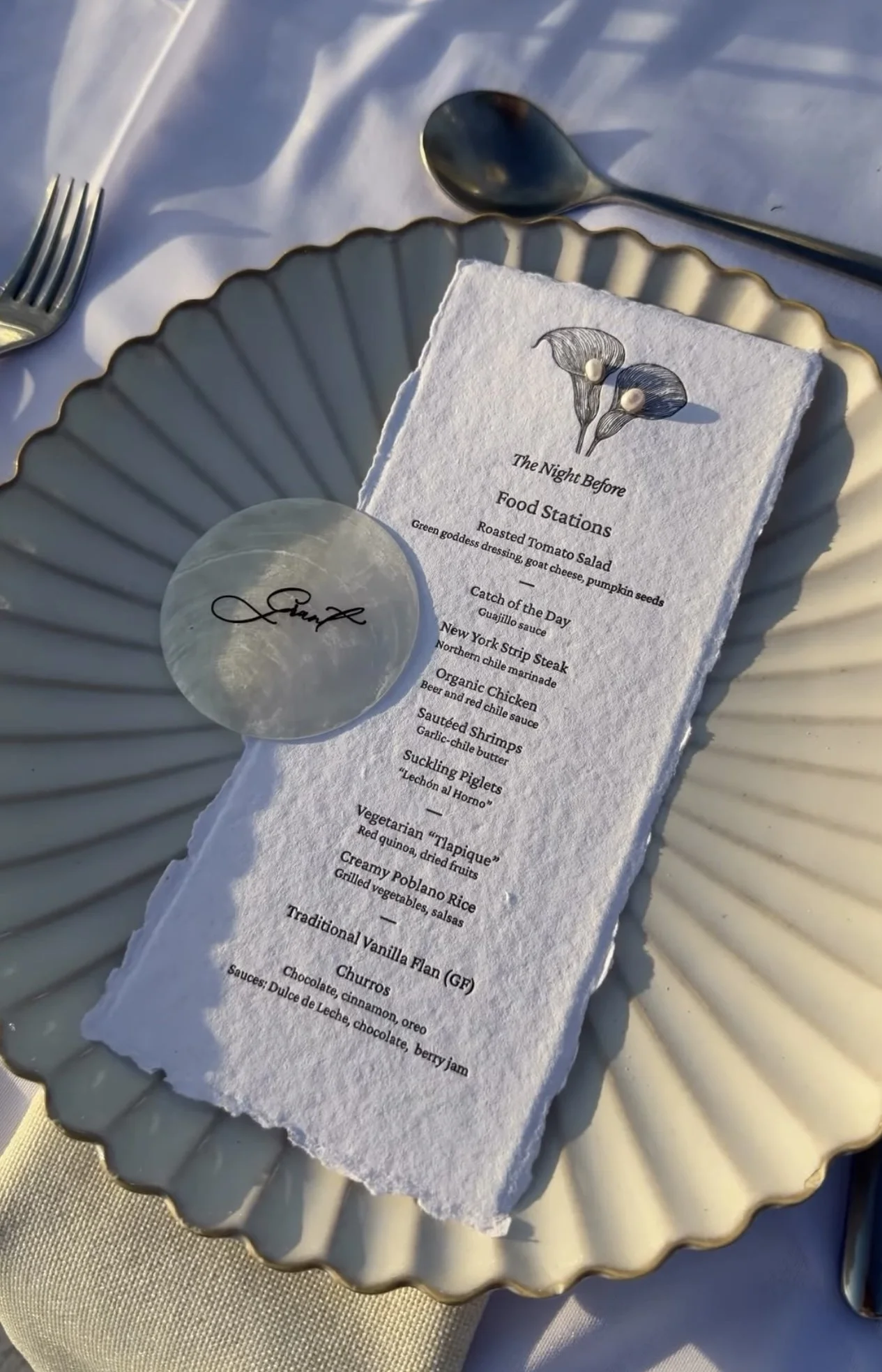 A table setting with a decorative plate, a menu titled 'The Night Before Food Stations,' and a garnet. The menu lists various food items such as roasted tomato salad, catch of the day, New York strip steak, organic chicken, sautéed shrimps, suckling piglets, vegetarian 'tapiquie,' creamy poblano rice, a traditional vanilla flan, and churros. The table is covered with a white cloth, and silverware is present.