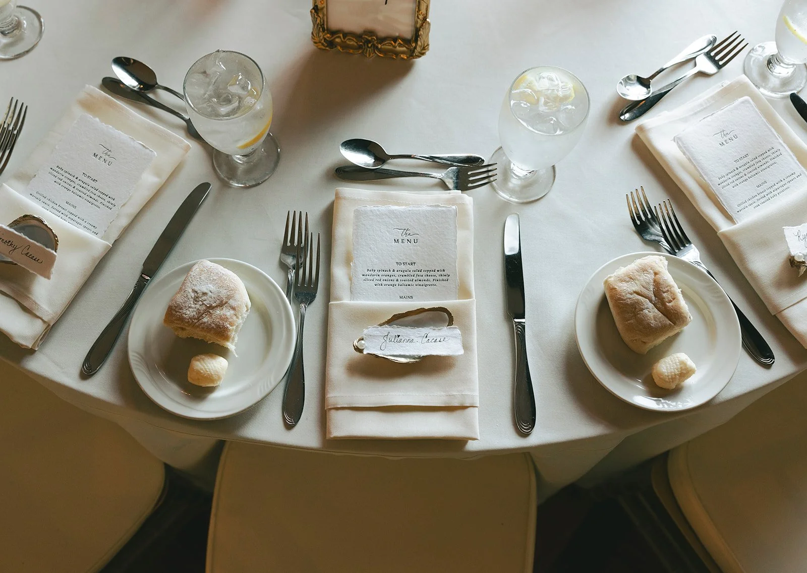 Elegant table setting with two place settings, each with a plate of bread, a small butter pat, a knife, a fork, a spoon, a napkin with a menu, and a glass of water with lemon, arranged on a white tablecloth.