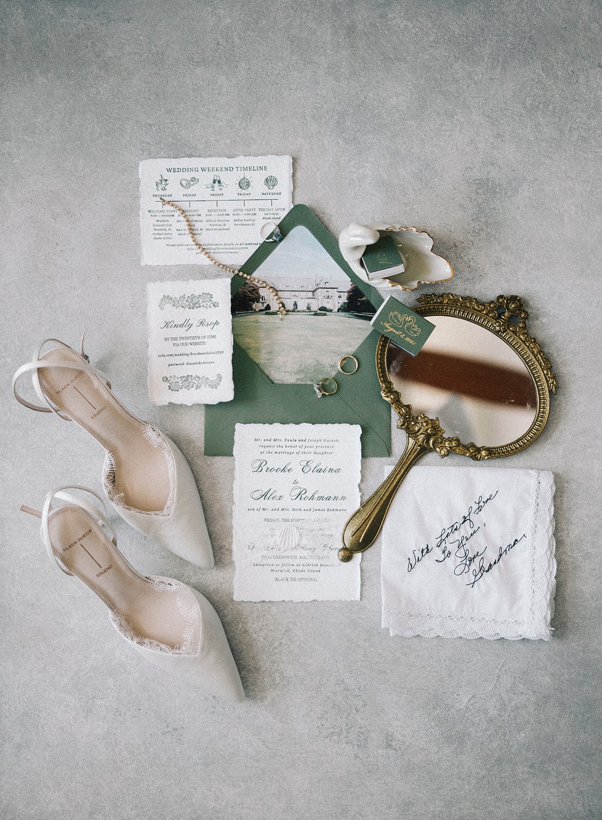 Bridal accessories and invitations arranged on a light surface, including white lace shoes, a vintage hand mirror, wedding invitations, a green envelope, a photograph, jewelry, a shell-shaped dish, and a handkerchief with a handwritten note.