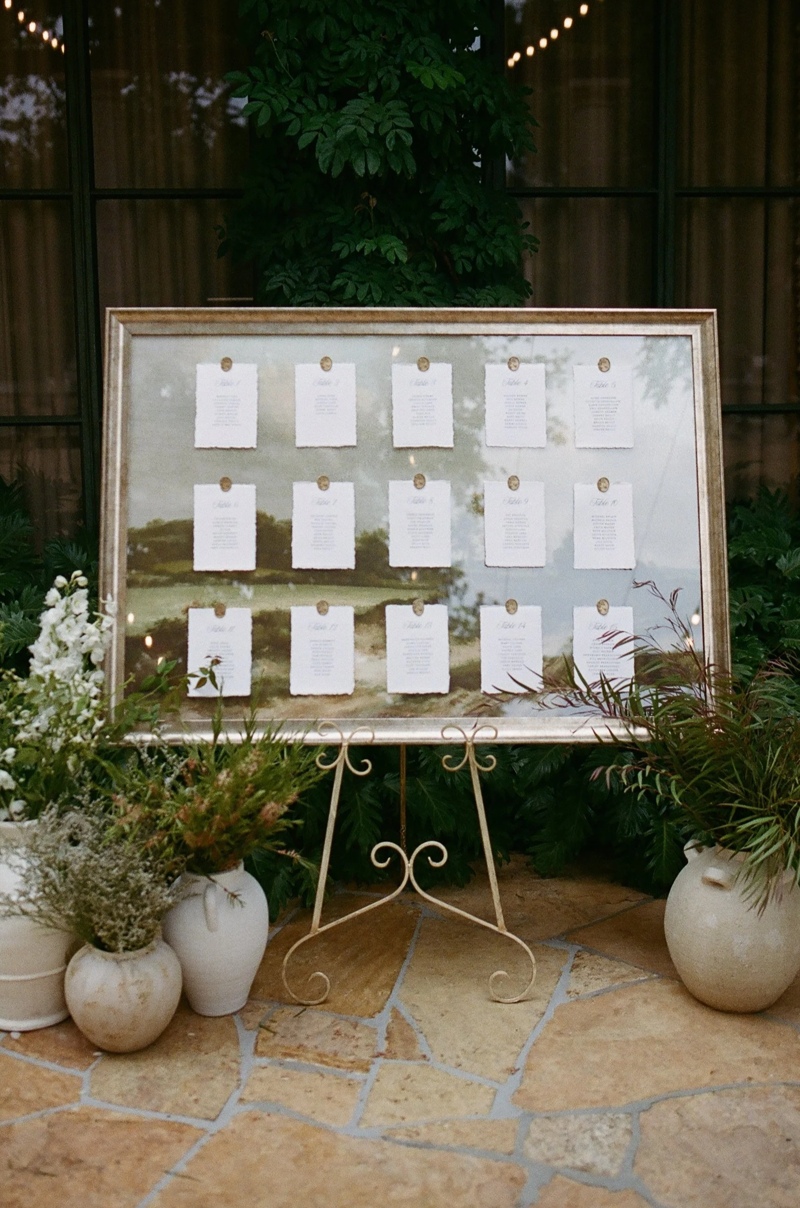 A wedding seating chart displayed on a mirror with a gold frame, surrounded by potted plants and flowers, situated on a tiled floor outside a building.